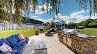 Located at Rockingham Court in Westerville, Ohio, this well-appointed outdoor community amenity area offers a thoughtfully designed space for recreation and relaxation. At its centerpiece is a large, multi-level play structure finished in blue and white, complete with tube slides, a yellow slide, a tunnel, climbing elements, and elevated walkways with decorative fencing — all set on a rubber mulch surface bordered by neat landscape edging for safety and visual appeal. Adjacent to the play structure, a dedicated swing set area features both toddler bucket seats and standard swings, providing options for children of all ages.

Beyond the play area, the grounds transition into a welcoming social space shaded by a graceful weeping willow tree. A stone fire pit surrounded by seating anchors this gathering zone, complemented by a wooden pergola with a picnic table and built-in grill — ideal for outdoor entertaining. Blue Adirondack-style benches are positioned throughout, offering relaxed seating with views of the lush green lawn.

Manicured landscaping with flowering hydrangeas, shrubs, and mulched garden beds lines the perimeter, while an American flag and decorative lamp post add a classic, community-oriented character. The overall ambiance is inviting, family-friendly, and thoughtfully maintained.