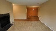 This single-floor space is thoughtfully divided into distinct functional zones, creating a sense of purposeful layout despite its open feel. The living area is finished with wall-to-wall carpet in a neutral beige tone, providing a soft, comfortable foundation that contrasts with the adjacent dining area. A step up transitions the space into a raised dining nook, finished with rich, dark hardwood-style flooring that adds warmth and visual distinction. This elevated area features a bold terracotta-orange accent wall, which serves as a striking focal point and adds depth to the otherwise neutral color palette. A single pendant light hangs centrally above the dining area, offering both functional illumination and a decorative touch. The surrounding walls throughout the space are finished in a light beige tone, contributing to a warm, cohesive ambiance. White baseboards provide clean contrast along the floor transitions. A ceiling vent is visible, indicating central climate control. The open sightlines between the two zones foster a connected feel while the flooring and color changes subtly define each area's purpose.