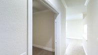 This single-floor space presents a clean, understated interior defined by neutral wall tones and classic white architectural trim. The hallway leading to an adjoining room showcases white-painted door framing accented by crown molding, adding a polished, traditional character to the space. The walls carry a warm beige palette, creating a calm and cohesive atmosphere throughout. Carpet flooring runs through the rooms, offering a soft underfoot feel, while the transition between spaces is marked by clean, well-maintained baseboards. The textured wall finish visible along the corridor adds subtle depth to the interior surfaces. Natural light appears to filter in through a window visible in the adjacent room, contributing to the overall brightness of the space. The layout suggests a straightforward, functional floor plan with clearly defined rooms connected by a central corridor. Crown molding is consistently applied, reinforcing the traditional aesthetic. Overall, the space maintains a simple, move-in-ready character suited to a variety of interior styles.