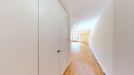 This single-floor residence presents a cohesive and functional layout defined by warm parquet flooring laid in a distinctive basket-weave pattern throughout the main living areas and hallway. The entryway corridor leads from a large built-in closet toward an open living space, where natural light streams through expansive windows, brightening the neutral-toned walls and highlighting the richly patterned wood floors.

The spacious main living area offers a generous, open feel with clean white ceilings and soft, warm wall tones. Multiple doorways connect the living space to the kitchen and additional rooms, creating a practical flow throughout the unit.

The kitchen is equipped with ample white cabinetry — both upper and lower — complemented by dark granite countertops that provide strong visual contrast. White appliances, including a gas range, dishwasher, microwave, and refrigerator, are neatly integrated within the L-shaped layout. Light tile flooring completes the kitchen's clean, utilitarian aesthetic.

The bathroom features a tiled tub-shower combination with a curtain rod, a warm-toned vanity with a granite countertop, and tile flooring, all set against cream-colored walls. The overall ambiance of the space is understated and well-maintained, with consistent design elements tying each area together cohesively.