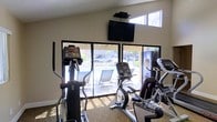 This fitness center is a compact yet functional workout space designed to support cardiovascular training and general exercise. The room is outfitted with multiple cardio machines, including elliptical trainers and a stationary recumbent bike, providing a variety of options for users at different fitness levels. A wall-mounted flat-screen television is positioned prominently above the equipment, offering entertainment during workouts. The space benefits from abundant natural light, courtesy of a row of large floor-to-ceiling windows along one wall. These windows not only brighten the interior but also provide an open, airy atmosphere and direct views of an outdoor patio area furnished with lounge chairs, creating a pleasant visual connection to the exterior. The neutral-toned walls and light carpeted flooring contribute to a clean and welcoming environment. Elevated window panels near the ceiling further enhance the flow of natural light throughout the room. The overall layout is straightforward and practical, making efficient use of the available floor space while ensuring ease of movement between equipment. This fitness center serves as a convenient and well-appointed amenity for those seeking an accessible space for their daily exercise routines.