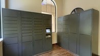 This package locker room combines modern convenience with classic architectural charm. The space is defined by its notably tall ceilings, which lend an open, airy quality to the room, and a prominent arched window that allows natural light to filter in, illuminating the warm, neutral-toned walls. The wood-style flooring adds a touch of warmth and complements the traditional aesthetic of the architecture.

The room is equipped with multiple rows of smart package lockers finished in a muted olive-gray tone, arranged strategically along the walls to maximize storage capacity. The lockers vary in compartment size, accommodating packages of different dimensions. A central locker unit features an integrated touchscreen kiosk with a keypad interface, enabling secure, self-service package retrieval. The system is designed to streamline package management, providing residents or users with a convenient and efficient solution for receiving deliveries at any time.

The arched architectural details visible on the walls echo the window design, reinforcing a cohesive, refined aesthetic throughout the space. The overall ambiance balances functional infrastructure with thoughtful design, making this package room both practical and visually appealing.