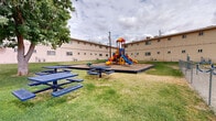 Located at 718 N Carolina Dr in El Paso, Texas, this residential complex presents a well-maintained, multi-building layout with a distinctive tan stucco exterior. The buildings span two stories, featuring evenly spaced windows throughout and individual HVAC units servicing each unit. The grounds surrounding the buildings are generously landscaped with maintained green lawns and mature shade trees that provide natural relief from the El Paso sun.

A central courtyard serves as the heart of the outdoor community space, anchored by a vibrant, multi-colored playground structure complete with slides, climbing elements, and a separate toddler play area, all set on a wood chip safety surface enclosed by timber edging. Additional outdoor amenities include multiple blue metal picnic tables positioned throughout the lawn areas, ideal for outdoor gatherings. A chain-link fenced area adjacent to the playground provides an additional recreational zone with swing-style fitness equipment.

Paved walkways run along the perimeter of the buildings, connecting residents to designated parking areas. A row of brown garage units along the property edge offers additional secure storage or covered parking options. The property's layout emphasizes communal outdoor living, making it a functional and family-friendly residential environment in the heart of El Paso.