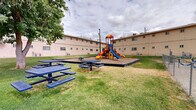 Located at 718 N Carolina Dr in El Paso, Texas, this residential complex presents a well-maintained, multi-building layout with a distinctive tan stucco exterior. The buildings span two stories, featuring evenly spaced windows throughout and individual HVAC units servicing each unit. The grounds surrounding the buildings are generously landscaped with maintained green lawns and mature shade trees that provide natural relief from the El Paso sun.

A central courtyard serves as the heart of the outdoor community space, anchored by a vibrant, multi-colored playground structure complete with slides, climbing elements, and a separate toddler play area, all set on a wood chip safety surface enclosed by timber edging. Additional outdoor amenities include multiple blue metal picnic tables positioned throughout the lawn areas, ideal for outdoor gatherings. A chain-link fenced area adjacent to the playground provides an additional recreational zone with swing-style fitness equipment.

Paved walkways run along the perimeter of the buildings, connecting residents to designated parking areas. A row of brown garage units along the property edge offers additional secure storage or covered parking options. The property's layout emphasizes communal outdoor living, making it a functional and family-friendly residential environment in the heart of El Paso.