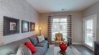 Located at 5260 Franklin St in Hilliard, Ohio, this single-floor residence combines modern finishes with inviting design details throughout. The living room serves as the centerpiece of the home, highlighted by a striking whitewashed brick accent wall, wood-look flooring, and large windows dressed in buffalo-check curtains that allow ample natural light to fill the space. The kitchen is outfitted with dark espresso cabinetry, stainless steel appliances including a refrigerator, microwave, and electric range, and a light stone-finish countertop that extends to a peninsula breakfast bar with pendant lighting overhead. Wood-look flooring continues seamlessly through the kitchen and common areas. The two bedrooms feature plush carpeting, generous natural light from large windows, and neutral wall tones that create a calm, restful atmosphere. Both bathrooms are finished with the same rich dark cabinetry, stone-topped vanities, large framed mirrors, and wood-look tile flooring. The primary bathroom includes a full tub-shower combination, while the second bathroom offers an equally refined design. Cohesive neutral tones, quality fixtures, and thoughtful design details are consistent throughout, giving the entire residence a polished and comfortable feel.