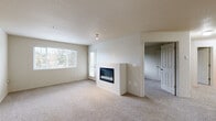 This single-floor residence offers a well-organized layout encompassing three bedrooms, two bathrooms, a combined kitchen and living area, a dedicated laundry room, a hallway, and a closet. The living room serves as the central gathering space, finished with neutral-toned carpet and anchored by a sleek, wall-mounted electric fireplace. Large windows allow natural light to fill the room while providing views of surrounding greenery, and a door offers direct exterior access. The kitchen features rich, dark wood cabinetry paired with dark granite countertops, stainless steel appliances including a refrigerator and range, and a raised breakfast bar that visually connects the kitchen to the living area. The bedrooms are carpeted and finished with neutral walls; each benefits from natural light, with at least one room including a baseboard heater beneath a window overlooking trees. The primary bedroom connects to a spacious walk-in closet outfitted with wire shelving for organized storage. The bathrooms include a full tub-shower combination with clean white surround. A dedicated laundry room houses a full-size washer and dryer with overhead wire shelving. Textured ceilings are consistent throughout, lending a uniform, cohesive finish to the space.