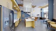 This contemporary single-floor space makes a striking impression with its dramatic double-height ceilings and open-plan layout that encourages fluid movement between functional zones. The kitchen is a standout feature, outfitted with warm yellow cabinetry, clean white countertops, and a curved island with built-in cooktop and bar seating. A bold stainless steel range hood serves as an architectural focal point, suspended from above and drawing the eye upward toward the soaring ceiling. Recessed lighting and a series of elegant pendant lights illuminate the space with layered warmth. Open shelving and a dedicated sink area along the perimeter wall add practical utility without compromising the open feel. The adjacent lounge area transitions naturally from the kitchen, furnished with modern seating and anchored by a wall-mounted display. A staircase with sleek metal railings adds vertical interest and contributes to the airy, multi-dimensional character of the interior. Blue accent walls punctuate the predominantly neutral palette, adding depth and visual energy. Large-format tile flooring and wood-paneled ceiling accents complete the sophisticated, design-forward aesthetic of this well-appointed space.