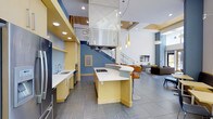 This contemporary single-floor space makes a striking impression with its dramatic double-height ceilings and open-plan layout that encourages fluid movement between functional zones. The kitchen is a standout feature, outfitted with warm yellow cabinetry, clean white countertops, and a curved island with built-in cooktop and bar seating. A bold stainless steel range hood serves as an architectural focal point, suspended from above and drawing the eye upward toward the soaring ceiling. Recessed lighting and a series of elegant pendant lights illuminate the space with layered warmth. Open shelving and a dedicated sink area along the perimeter wall add practical utility without compromising the open feel. The adjacent lounge area transitions naturally from the kitchen, furnished with modern seating and anchored by a wall-mounted display. A staircase with sleek metal railings adds vertical interest and contributes to the airy, multi-dimensional character of the interior. Blue accent walls punctuate the predominantly neutral palette, adding depth and visual energy. Large-format tile flooring and wood-paneled ceiling accents complete the sophisticated, design-forward aesthetic of this well-appointed space.
