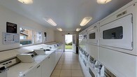 This single-floor communal laundry facility is designed for efficiency and ease of use. The room is laid out in a long, corridor-style format that maximizes the use of available space. Along the left wall, a generous row of top-loading washing machines is arranged in sequence, providing ample capacity for multiple users. On the right side, stacked dryer units line the wall from floor to near-ceiling, offering a substantial number of drying options to accommodate high-volume laundry needs. The floor is finished with light-toned ceramic tiles that are easy to clean and maintain, contributing to the room's bright and functional aesthetic. Overhead, flush-mounted fluorescent light fixtures run the length of the ceiling, ensuring the space is well-lit throughout. The walls are finished in a neutral off-white tone, keeping the environment clean and simple. At the far end of the room, a white-framed glass door opens to an outdoor area, allowing natural light to filter in and providing ventilation. A window along the left wall further enhances the natural brightness of the space. Overall, the facility is practical, spacious, and well-maintained.