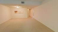 This single-floor space offers a clean, open layout designed for comfortable everyday living. The main living area is generously sized, featuring wall-to-wall neutral-toned carpeting that lends warmth and continuity throughout the space. The walls are finished in a soft, light color palette, contributing to a bright and airy atmosphere even without abundant natural light.

A notable architectural feature is the pass-through window connecting the living area to the kitchen, allowing for convenient interaction between the two spaces. The kitchen, visible through the pass-through, includes a sink and cabinetry, offering a practical and efficient workspace. Overhead lighting in the living area provides consistent illumination, while a ceiling vent indicates a central HVAC system for climate control.

A hallway leading from the main living area provides access to additional rooms, suggesting a well-organized floor plan. Doorways visible along the corridor hint at further private spaces beyond the main living zone. Overall, the space presents a neutral, move-in-ready environment with a straightforward layout suited to a variety of living arrangements.