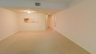 This single-floor space offers a clean, open layout designed for comfortable everyday living. The main living area is generously sized, featuring wall-to-wall neutral-toned carpeting that lends warmth and continuity throughout the space. The walls are finished in a soft, light color palette, contributing to a bright and airy atmosphere even without abundant natural light.

A notable architectural feature is the pass-through window connecting the living area to the kitchen, allowing for convenient interaction between the two spaces. The kitchen, visible through the pass-through, includes a sink and cabinetry, offering a practical and efficient workspace. Overhead lighting in the living area provides consistent illumination, while a ceiling vent indicates a central HVAC system for climate control.

A hallway leading from the main living area provides access to additional rooms, suggesting a well-organized floor plan. Doorways visible along the corridor hint at further private spaces beyond the main living zone. Overall, the space presents a neutral, move-in-ready environment with a straightforward layout suited to a variety of living arrangements.