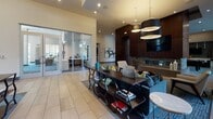 This thoughtfully designed amenity space presents a seamless blend of contemporary style and functional comfort across a single level. The grand lobby sets an impressive tone with soaring floor-to-ceiling windows that flood the interior with natural light and frame views of a lush, landscaped courtyard surrounded by the residential community.

The main lounge area is generously proportioned, featuring large-format tile flooring, recessed lighting, and a distinctive coffered wood ceiling detail with warm ambient uplighting. Multiple seating arrangements — including plush sofas and accent chairs in rich blues and grays — create inviting spaces for relaxation or socializing, anchored by a wall-mounted television and tasteful artwork throughout.

A dedicated game room offers leisure amenities including a billiard table, foosball table, and a sleek fireplace set against a richly paneled wood wall, providing a sophisticated yet playful retreat. Adjacent to the lounge, a fully appointed kitchen and bar area showcases an expansive marble-topped island with bar seating, herringbone tile backsplash, dark wood cabinetry, and pendant lighting — ideal for entertaining.

The mailroom and entry corridor feature eye-catching geometric patterned carpet, elegant pendant fixtures, and curated wall art, ensuring a polished first impression. Glass-walled office or co-working spaces add a practical dimension to this well-rounded communal facility.