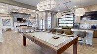Located at Vintage Brunswick in Brunswick, Georgia, this thoughtfully designed single-floor clubhouse offers a generously sized community gathering space that blends modern coastal aesthetics with functional amenities. The entry foyer welcomes visitors with striking chevron-patterned hardwood floors, soaring coffered ceilings, white shiplap-clad columns, and an elegant chandelier that sets a sophisticated tone throughout.

The expansive main living area serves as the heart of the space, anchored by a dramatic floor-to-ceiling tiled fireplace wall with an integrated linear electric fireplace and a mounted flat-screen TV above. Natural light pours in through large windows dressed with neutral drapery, creating a bright and inviting atmosphere. A billiards table takes center stage in the lounge area, complemented by statement pendant lighting and a cozy seating arrangement nearby.

The kitchen and bar area features warm wood cabinetry, quartz countertops, beverage refrigerators, and multiple coffee and drink stations, all adjacent to an industrial-style glass garage door that opens to the outdoors. A dedicated dining island with upholstered bar seating rounds out this entertaining zone.

The office suite offers a quiet retreat with a built-in dual workstation, soft carpeting, sage-toned accent walls, and decorative wall art, with an adjacent glass-walled conference room providing additional collaborative workspace.