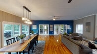 This single-floor residence showcases a thoughtfully designed open-concept layout that seamlessly connects the living, dining, and kitchen areas. Rich hardwood floors run throughout the main living spaces, complemented by bold navy blue accent walls that lend a striking, cohesive character to the interior. Vertical shiplap-style wall paneling in a warm gray tone adds texture and depth, while crown molding provides a refined finishing touch.

The living room is anchored by a classic white brick fireplace with a wood mantel, creating a cozy focal point. Ceiling fans maintain comfort throughout the open areas. French doors connect various spaces, enhancing the flow and openness of the floor plan.

The dining area is a standout space, flooded with natural light through expansive sliding glass doors that open directly onto a spacious outdoor deck. A live-edge wooden dining table sits beneath a modern cluster pendant light fixture, and the surrounding views of lush greenery create a serene atmosphere.

The kitchen features white shaker-style cabinetry, a mosaic tile backsplash, dark countertops, and a peninsula with bar stool seating — ideal for casual dining. Black appliances provide a bold contrast to the bright cabinetry.

A welcoming sitting area near the entryway, with double doors opening to a tiled foyer, rounds out this well-appointed and visually dynamic single-floor home.