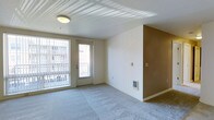This single-floor unit offers a clean, functional layout designed for comfortable everyday living. The main living area is a standout feature, characterized by plush wall-to-wall carpeting that adds warmth underfoot and a textured ceiling that lends subtle visual interest overhead. A large window paired with a glass-paneled exterior door floods the room with natural light and provides direct access to a private balcony, where views of the surrounding building exterior can be enjoyed. The neutral white walls are complemented by a warm tan accent wall, creating a welcoming sense of depth and dimension in the space. A wall-mounted thermostat and a built-in heating unit ensure year-round climate control. The overall aesthetic is understated and versatile, offering a blank canvas well-suited for a variety of interior styles. The unit's straightforward single-floor design ensures easy navigation throughout, making it a practical and accessible living space. Natural light plays a central role in the ambiance, keeping the interior feeling open and airy throughout the day.