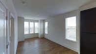 This single-floor space presents a clean, modern aesthetic defined by its crisp white walls and smooth ceilings. The main living area is anchored by warm hardwood-style flooring that runs throughout, lending a sense of continuity and warmth to the interior. Multiple large windows fitted with horizontal blinds line the walls, including a corner configuration that draws in generous amounts of natural light from multiple angles. These windows also frame views of the surrounding exterior, enhancing the sense of openness and connectivity to the outdoors. The layout is open and unobstructed, offering a versatile floor plan that can accommodate a variety of uses and furnishing arrangements. Recessed lighting in the ceiling provides supplemental illumination, complementing the abundant daylight. White baseboards provide a clean finishing touch along the perimeter of the room, contributing to the overall polished and well-maintained appearance of the space. The overall ambiance is bright, airy, and move-in ready, with a neutral palette that serves as an ideal canvas for personalization.