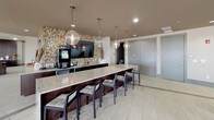 Located at 1316 E 1st St in Charlotte, NC, this single-floor amenity space is designed to serve as a sophisticated social and recreational hub. The interior features large-format light tile flooring throughout, dark wood trim accents, and recessed lighting that complement the contemporary aesthetic.

The centerpiece of the space is a well-appointed bar and kitchen area, anchored by an elongated marble-look quartz countertop with dark wood cabinetry below. Globe pendant lights hang above the bar seating, while a striking mosaic tile accent wall frames a wall-mounted television, adding a vibrant focal point. Adjacent kitchen appliances and ample counter space make the area well-suited for entertaining.

The lounge zone features comfortable upholstered seating arranged around low tables, with large floor-to-ceiling accordion glass doors that open directly onto an outdoor terrace. The terrace offers views of the Charlotte skyline and is furnished with vibrant seating for outdoor gatherings. Clerestory windows above the accordion doors invite abundant natural light into the interior.

A co-working area provides high-top bistro tables positioned alongside large windows overlooking the city. A billiards table occupies a central area of the room, accompanied by curtain-framed windows. A separate, more intimate lounge nook features oversized windows with draped curtains and views of the surrounding neighborhood, rounding out this multifunctional community space.