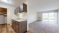This single-floor unit offers a practical and comfortable layout with distinct living, kitchen, and bathroom areas. The galley-style kitchen is equipped with dark wood cabinetry on both upper and lower levels, a double sink with light-toned countertops, a white electric range, a refrigerator, and durable wood-look vinyl flooring. An adjacent open area connects seamlessly to the kitchen, providing a flexible dining or additional living space.

The living area is generously sized and finished with neutral wall-to-wall carpeting, contributing to a warm and quiet atmosphere. A sliding glass door fills the room with natural light and provides outdoor views of surrounding greenery, while a wall-mounted air conditioning unit offers convenient climate control. Baseboard heating runs along the perimeter of the main living spaces and bedrooms.

The bedrooms are carpeted and well-lit by large windows that frame outdoor views, including what appears to be a scenic water feature from one room. The bathroom features a full-length tub and shower combination, a marble-patterned vanity countertop with an integrated sink, a large mirror, and classic mosaic tile flooring. A walk-in closet provides additional storage near the kitchen area.