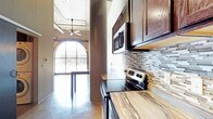This single-floor space showcases an industrial loft aesthetic, thoughtfully balanced with contemporary updates throughout. The open living area immediately impresses with soaring ceilings, polished concrete floors that reflect natural light, and exposed structural columns that serve as architectural focal points. Ceiling fans circulate air through the generous vertical space, enhancing the airy, open feel.

A striking arched window framed by original brick masonry anchors one end of the living area, flooding the space with natural light and offering unobstructed views of the surrounding streetscape. Exposed ductwork and industrial-style track lighting overhead reinforce the loft character.

The kitchen is well-appointed with rich, dark-stained wood cabinetry extending to the upper reaches of the tall walls, providing ample storage. A mosaic tile backsplash in neutral tones of gray, white, and beige adds texture and visual interest. Stainless steel appliances—including a range, microwave, dishwasher, and refrigerator—complement the marble-veined countertops, striking a balance between industrial and refined.

The bathroom continues the space's consistent design language, featuring a dark wood vanity with a stone-top countertop, a large mirror, and a walk-in shower with clean white surround panels.