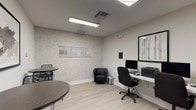 This single-floor business center offers a well-organized and functional environment designed to support productivity and collaboration. The space is finished in a clean, neutral palette of soft grays and whites, complemented by light wood-look flooring that runs throughout, lending a cohesive and professional feel.

The workstation area is equipped with multiple desktop computers and a printer arranged along a wall-mounted white desk, paired with ergonomic black office chairs. A framed abstract artwork hangs above the workstation, adding a refined touch to the otherwise utilitarian setup. A black leather accent chair provides a comfortable secondary seating option within the same area.

Adjacent to the workstation zone is a casual lounge and meeting area furnished with round bistro-height tables and modern black slatted bar stools — ideal for informal gatherings or quick discussions. A large landscape-style framed print decorates the wall, enhancing the ambiance of the space.

The rooms are accessed through a classic white French door with grid-pane glass panels, connecting the spaces while maintaining an open sightline to a larger adjoining area visible in the background. Recessed fluorescent ceiling fixtures provide consistent, even lighting throughout. Small windows fitted with horizontal blinds allow for natural light control. The overall design is minimal, functional, and polished, making it well-suited for a shared office or community business center setting.