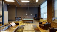 This single-floor space exudes an industrial-chic aesthetic, blending raw architectural character with comfortable, social amenities. The centerpiece of the room is a full-size billiards table, elegantly lit by a sleek, multi-pendant linear chandelier that draws the eye upward toward the striking reclaimed wood plank ceiling. Exposed brick walls and dark, charcoal-painted surfaces create a moody, urban atmosphere, while wide-plank hardwood floors add warmth and texture throughout. Natural light filters in generously through large windows along one wall, brightening the space and highlighting its rich tones. Leather sofas and a patterned area rug define a relaxed lounge area, perfect for gathering around the game. High-top bar tables with stools are positioned in the corners, offering additional seating for socializing. Visible ductwork and structural elements complement the loft-style design, reinforcing the space's industrial character. The overall ambiance is inviting yet bold — a well-appointed retreat designed for entertainment and leisure.