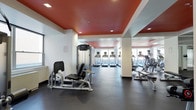 This fitness center offers a thoughtfully designed workout environment spread across a single level. The space is immediately distinguished by its striking red-painted ceiling panels, which contrast sharply with the clean white walls and structural columns, creating an energetic and motivating atmosphere. Recessed lighting is evenly distributed throughout, ensuring the entire area is well-lit for safe and comfortable exercise. The flooring consists of dark rubber matting, a practical and durable surface well-suited for heavy equipment and high-traffic use. On one side of the space, a row of treadmills by Precor lines the perimeter, each positioned facing windows that allow natural light to filter in, providing an open and airy feel during cardio sessions. Opposite, additional cardio equipment including elliptical machines and steppers rounds out the aerobic offerings. Strength training is supported by cable resistance machines positioned toward the front of the room. Structural columns divide the space into distinct workout zones without disrupting the overall flow. The combination of bold design elements, quality equipment, and natural light makes this fitness center a functional and visually engaging space.