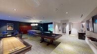 This sports lounge in Spring, Texas, is a thoughtfully designed entertainment space that brings together a diverse array of recreational amenities under one roof. The open main area is anchored by a full-size billiards table with a dark felt surface, complemented by a wall-mounted cue rack, and a shuffleboard table positioned nearby for casual play. A dartboard adds another classic option for guests seeking friendly competition. The centerpiece of the lounge is a state-of-the-art golf simulator, set within a dedicated alcove featuring a projection screen, putting surface, and a monitor for tracking play — all framed by dark, dramatic walls that enhance the immersive experience. Distinctive globe pendant lighting and recessed fixtures illuminate the space with a warm, layered glow, while a patterned mosaic-style carpet adds visual interest underfoot. A dedicated media nook features a large wall-mounted flat-screen TV surrounded by plush, textured lounge chairs and small side tables, creating a comfortable spot for watching sports or films. A row of bold red storage lockers adds a pop of color to the locker area. The wet bar corridor is finished with granite countertops, an undermount sink, under-counter cabinetry, wall sconces, and its own mounted display screen, providing a convenient refreshment station for the entire lounge.