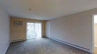 This single-floor residence offers a straightforward, functional layout designed for comfortable everyday living. The main living area is finished with wall-to-wall carpet in a neutral tone, providing a soft underfoot feel throughout the space. Warm beige walls contribute to a calm and inviting atmosphere, while the standard flat ceiling keeps the room feeling clean and uncluttered. A prominent sliding glass door fitted with vertical blinds serves as both a source of natural light and direct access to an outdoor balcony or patio area, offering a pleasant connection to the exterior. A wall-mounted air conditioning unit positioned near the sliding door ensures climate control during warmer months, while baseboard heaters running along the perimeter of the room provide efficient warmth during cooler seasons. An interior door adjacent to the sliding glass access point leads to additional areas of the unit. The overall design is practical and neutral, presenting a blank canvas that accommodates a variety of furnishing styles and personal preferences. The space is well-suited for those seeking a low-maintenance, single-level living environment with essential amenities already in place.