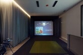 This contemporary indoor golf simulator facility is thoughtfully designed to deliver an immersive, high-tech sporting experience. The centerpiece of the space is a Full Swing golf simulator bay, featuring a large high-definition projection screen, a ceiling-mounted projector, and a professional artificial turf hitting mat. Floor-to-ceiling dark paneling frames the simulator enclosure, creating a focused and distraction-free environment for players of all skill levels.

Adjacent to the simulator bay, a comfortable lounge area provides space for guests to relax and socialize. High-top bar tables paired with cushioned bar stools offer casual seating, while soft accent lighting installed along the ceiling perimeter creates a warm, intimate ambiance throughout. Floor-length neutral-toned drapes line the walls, contributing to the refined and polished aesthetic of the space.

A reception counter separates the lounge from the simulator, providing a practical service point and an organizational hub for the facility. Beyond the counter, a media wall featuring a flat-screen television and cabinetry offers additional entertainment options. Light wood-tone flooring runs through the lounge and reception areas, contrasting elegantly with the dark carpeting of the simulator zone. Glass-panel entry doors and interior windows allow for visibility while maintaining a clean, modern architectural character throughout.