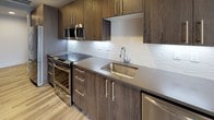 This contemporary single-floor unit showcases a clean, modern design throughout, with light gray walls, smooth recessed ceilings, and light-toned wood-style flooring that flows seamlessly across the main living areas. The open layout offers a generous living space with ample room for various furniture configurations, complemented by large floor-to-ceiling windows that frame elevated urban views of the surrounding cityscape, filling the interior with natural light.

The kitchen is thoughtfully designed with rich, dark wood cabinetry, gray quartz countertops, a textured white tile backsplash, and a full suite of stainless steel appliances including a range, microwave, and dishwasher. Dark floating shelves and a built-in shelving unit provide additional display and storage options adjacent to the main living area.

The bedroom features rich, wide-plank hardwood flooring and a striking floor-to-ceiling built-in wardrobe finished in dark gray wood grain with sleek silver hardware, offering substantial and well-organized storage.

The bathroom is a standout space, appointed with a large glass-enclosed walk-in shower clad in elegant marble-style tile, hexagonal mosaic floor tile, and wood-toned large-format floor tiles. A wide vanity with warm wood cabinetry, a gray stone countertop, an under-mount sink, and a backlit mirror complete the spa-like aesthetic. A ceiling fan adds comfort to the bedroom area, which connects directly to the bathroom.