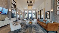 This single-floor communal lounge and lobby space is designed to impress with its soaring double-height ceilings and expansive, open layout that conveys a grand sense of arrival. The interior is anchored by rich wood-plank flooring that transitions seamlessly into multi-toned area rugs, grounding the carefully curated seating arrangements throughout. Statement geometric pendant chandeliers hang from the lofty ceiling, complemented by wall-mounted sconces that add warm, layered lighting across the space.

The lounge area is furnished with a mix of a classic cognac leather sofa, upholstered accent chairs, and sleek wooden side tables, striking a balance between comfort and contemporary style. Large black-framed windows and glass doors flood the interior with abundant natural light while offering views of the outdoor surroundings. A standout herringbone-patterned brick accent wall adds texture and visual depth, paired with a gallery wall of framed black-and-white artwork that elevates the aesthetic further.

Concrete-finished columns punctuate the open floor plan, reinforcing the modern-industrial character of the space. Multiple glass-panel doors lead to adjacent rooms and corridors, suggesting a well-connected and functional layout. Overall, the space exudes a refined yet approachable ambiance suited for gathering, socializing, or relaxing.