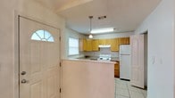 Located at 7791 Lilah Ln in Pensacola, Florida, this single-story home offers a straightforward and functional layout suited for comfortable everyday living. The kitchen is a central feature of the home, showcasing light wood cabinetry that provides ample storage space. A half-wall peninsula separates the kitchen from the adjacent area, creating a natural division while maintaining an open feel between spaces. The kitchen is equipped with a white electric range and a full-size white refrigerator, complemented by a under-cabinet range hood. Pendant lighting hangs above the peninsula, adding a subtle decorative touch. Light tile flooring runs throughout the space, offering both durability and ease of maintenance. A window above the kitchen sink allows natural light to filter into the cooking area. The entryway is highlighted by a classic white front door featuring a decorative arched transom window, welcoming natural light into the home's entry point. The textured ceiling adds a traditional finish consistent throughout the space. Overall, this home presents a clean, practical interior with a warm and approachable character well-suited to the Pensacola, Florida lifestyle.