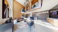 This thoughtfully designed single-floor space on Stadium Drive in Kansas City, Kansas, serves as a striking communal lobby and lounge area. The space is defined by its soaring double-height ceilings, which create an expansive, airy atmosphere and are anchored visually by an upper-level mezzanine with a metal railing, clad in warm wood paneling. Suspended woven pendant light fixtures add an artisan touch overhead.

The living area features a curved, sculptural sofa in a warm taupe tone, paired with contemporary accent chairs and a round pedestal coffee table, all arranged atop a large oval area rug with fluid, marble-like patterning. Polished concrete floors extend throughout, lending an industrial-modern aesthetic that contrasts beautifully with the natural wood wall cladding.

A reception desk anchors one end of the space, set against a full dark brick accent wall bearing bold signage. Floor-to-ceiling glazed entry doors flood the hallway with natural light and provide clear outdoor views. Additional brick detailing, lush potted greenery, and curated artwork contribute to the welcoming, community-forward character of this well-appointed space.
