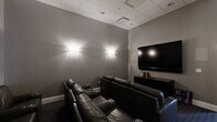 This purpose-built screening room offers a well-appointed home theater experience within a compact, efficiently designed single-floor layout. The space is accessed through a gracefully curved entry corridor, finished with textured grey wallcovering, dark patterned carpeting, and soft wall sconces that establish an immediate cinematic ambiance.

The main viewing area is designed with an emphasis on comfort and functionality. Multiple rows of plush, dark leather reclining seats — each equipped with built-in cup holders and armrests — face a large wall-mounted flat-screen television, supported by a low black media console. A small media library is organized neatly beside the console, adding a practical touch.

The room's interior is unified by consistent textured grey wall treatments and a drop ceiling with recessed panel tiles, contributing to a clean, modern aesthetic. Ambient wall sconces are symmetrically placed to provide soft, flattering light without disrupting the viewing environment. A structural column is incorporated seamlessly into the room's layout.

A glazed exit door at the rear connects the screening room to an adjacent corridor, ensuring easy accessibility. The overall design is sleek and purpose-driven, making this space a functional and inviting venue for private screenings or media entertainment.