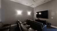 This purpose-built screening room offers a well-appointed home theater experience within a compact, efficiently designed single-floor layout. The space is accessed through a gracefully curved entry corridor, finished with textured grey wallcovering, dark patterned carpeting, and soft wall sconces that establish an immediate cinematic ambiance.

The main viewing area is designed with an emphasis on comfort and functionality. Multiple rows of plush, dark leather reclining seats — each equipped with built-in cup holders and armrests — face a large wall-mounted flat-screen television, supported by a low black media console. A small media library is organized neatly beside the console, adding a practical touch.

The room's interior is unified by consistent textured grey wall treatments and a drop ceiling with recessed panel tiles, contributing to a clean, modern aesthetic. Ambient wall sconces are symmetrically placed to provide soft, flattering light without disrupting the viewing environment. A structural column is incorporated seamlessly into the room's layout.

A glazed exit door at the rear connects the screening room to an adjacent corridor, ensuring easy accessibility. The overall design is sleek and purpose-driven, making this space a functional and inviting venue for private screenings or media entertainment.