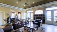 This inviting single-floor space is designed with both comfort and style in mind. The lounge area immediately draws attention with its distinctive tray ceiling, accented by recessed lighting and a sleek ceiling fan that enhances the room's airy, open feel. Warm, neutral wall tones complement the rich, dark-stained hardwood-style flooring and the plush area rug that anchors the seating arrangement.

The main gathering area is furnished with generous dark leather sofas and armchairs, arranged around a wooden coffee table to create an ideal setting for conversation or relaxation. A prominent fireplace serves as a focal point on one wall, flanked by built-in shelving units finished in dark tones, adding both functionality and architectural character.

Natural light flows generously into the space through multiple windows, including a striking arched transom window above the entryway that adds a refined architectural touch. French doors provide direct access to the outdoors, further brightening the interior. A small dining or refreshment area is visible in the background, complete with pendant lighting, offering an additional functional zone within the space. Overall, the layout and finishes create a warm, polished ambiance suited for communal gathering and everyday use.