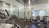 This single-floor fitness facility is purpose-built for effective strength and conditioning training. The space is defined by its clean, functional design, featuring corrugated metal wall paneling that gives the room an industrial character while remaining bright and open. Full-length mirrors line one wall, amplifying the sense of space and allowing users to monitor their form during exercise. The flooring throughout is durable rubber, providing both safety and comfort underfoot. At the center of the room stands a multi-functional cable machine, suitable for a wide range of resistance exercises. Flanking the space are organized dumbbell racks offering a full selection of free weights, paired with a dedicated weight bench for targeted strength work. Additional cardio and resistance machines are positioned throughout, making the facility well-suited for full-body training routines. A window near the entrance allows natural light to filter in, contributing to a more energizing workout environment. The overall ambiance is utilitarian yet well-maintained, offering users everything needed for a focused and productive training session.