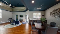 This single-story leasing office for Klingbeil Capital Management is designed to be both professional and welcoming. The interior is immediately distinguished by its dramatic tray ceilings, painted in a rich teal-blue tone that contrasts sharply with the neutral gray walls below, creating a visually striking architectural detail throughout the space. Warm hardwood flooring flows consistently across the entire floor plan, lending a cohesive and polished feel.

The main reception area greets visitors with a branded entry mat, teal-painted glass doors, and a comfortable seating arrangement featuring blue leather chairs and a low coffee table atop a geometric area rug. A wall-mounted flat-screen television and decorative wall art add character to the lounge. Natural light enters generously through multiple windows fitted with white blinds, offering views of surrounding greenery.

A dedicated workstation area with dark wood desks accommodates leasing staff, while a separate windowed office provides a quieter setting. The kitchenette features white quartz countertops, dark shaker cabinets, a stainless steel sink, and a self-serve coffee bar. Pendant lighting above the dining table and recessed ceiling lights complete the functional yet inviting atmosphere.