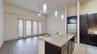 This single-floor residence offers a well-designed layout that balances functionality with modern finishes. The open-concept kitchen is a standout feature, showcasing rich dark cabinetry, granite countertops, a large center island with pendant lighting, stainless steel appliances including a refrigerator and range, and a tile backsplash. Tile flooring grounds the kitchen area, while hardwood-style flooring extends throughout the main living spaces, lending a cohesive and contemporary feel.

The living area is bright and airy, benefiting from large French doors and oversized windows fitted with blinds that invite abundant natural light. A ceiling fan anchors the space, and the elevated ceiling height adds a sense of openness. Bedrooms continue the hardwood-style flooring and feature large windows that allow natural light to fill the rooms.

A carpeted secondary space provides a softer area, ideal for flexible use. The bathroom is well-appointed with tile flooring, a tiled tub-shower surround, a granite-topped vanity with dark cabinetry, and clean, neutral finishes throughout.

Connecting the spaces is a hallway with track lighting and crisp white walls, ensuring smooth flow across the floor plan. Overall, the residence presents a clean, neutral palette with quality finishes at every turn.