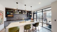 Located at 10 Calm Water Dr in Asheville, NC, this thoughtfully designed single-floor space seamlessly integrates a kitchen, dining room, and living room into one cohesive, open-plan layout. The kitchen serves as a central focal point, anchored by a waterfall-edge quartz island with seating for multiple guests, a gold-tone faucet, and a sleek undermount sink. Dark tile backsplash, open wood shelving, and light grey cabinetry line the back wall, while a trio of black-and-brass pendant lights adds a striking modern touch. Large black-framed floor-to-ceiling glass doors and windows flood the space with natural light and offer direct access to an outdoor deck with wooded views. The dining area continues the airy, light-filled atmosphere, featuring a minimalist wood dining table beneath a contemporary linear chandelier, all surrounded by expansive black-framed glass walls. The living room provides a warm contrast, centered around a linear electric fireplace set into a textured stone accent wall, complemented by a comfortable sectional sofa, built-in bench seating, and curated artwork. Large-format marble-look tile flooring unifies the entire space, while exposed ductwork adds an industrial edge to the modern aesthetic.