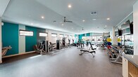 Located in Phoenix, Arizona, this fitness center offers a generously sized, single-floor exercise facility designed to accommodate a wide range of workout styles and fitness goals. The main exercise room is expansive and open, finished with durable rubber flooring and illuminated by recessed lighting complemented by ceiling fans that maintain a comfortable environment throughout. Bold teal accent walls punctuate the otherwise crisp white interior, lending the space a modern, energetic aesthetic.

The facility is comprehensively equipped with a full array of strength training machines, adjustable weight benches, and an extensive dumbbell rack suited for all fitness levels. Cardio equipment includes elliptical trainers, stationary bikes — including an air resistance bike — and additional machines positioned along a windowed wall that allows natural light to filter in. High-mounted horizontal windows line the perimeter, providing brightness without compromising wall space used for equipment.

A dedicated Training Zone area features a multi-functional rack stocked with kettlebells, medicine balls, resistance bands, and TRX suspension trainers, ideal for functional and group training. A separate hallway connects to additional storage and utility areas, including a styled accent wall with decorative tile. Full-length mirrors enhance the sense of space and support proper form during workouts. The overall layout is thoughtfully organized to ensure fluid movement and a focused, professional fitness environment.