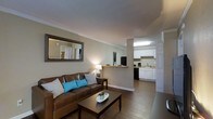 This 1-bedroom, 1-bathroom residence is thoughtfully designed to maximize comfort and functionality within a single-floor layout. The open-concept living and kitchen area creates a sense of spaciousness, with the living room serving as a warm and inviting central hub. Wood-tone flooring runs throughout, lending a cohesive and clean aesthetic to the space. Crown molding adds a refined architectural detail along the ceiling line, elevating the overall finish of the interior.

The living room is anchored by a rich brown leather sofa accented with teal and neutral throw pillows, paired with a dark wood coffee table. A framed wall mirror enhances the sense of depth and light in the room. A half-wall partition gracefully separates the living area from the kitchen, maintaining an open feel while defining each space.

The kitchen features white cabinetry with ample storage, a black appliance suite including a refrigerator and dishwasher, and a clean white countertop with a sink positioned along the far wall. The layout is efficient and well-organized, making it practical for everyday use. Overall, the space presents a functional, comfortable, and neatly finished interior well-suited for single occupancy or a couple.