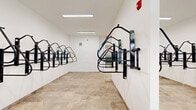 Located at 4021 Ridge Ave in the Roxborough neighborhood of Philadelphia, PA, this dedicated bicycle storage room offers a well-organized and functional space for residents to securely store their bikes. The room is laid out on a single floor and provides a generous, open floor plan that comfortably accommodates multiple wall-mounted bicycle racks installed along all interior walls, maximizing storage capacity while keeping the central floor area clear and accessible.

The space features neutral white walls that contribute to a clean, utilitarian aesthetic, while the stone tile flooring adds a durable and attractive finish underfoot. Fluorescent strip lighting runs along the ceiling, ensuring the room is well-lit throughout. A centrally positioned partial wall further expands the available rack mounting surface, effectively increasing storage without reducing usability.

Natural light filters in through a window at the far end of the room, offering a view of the surrounding exterior. A clearly marked exit door provides direct access to the outside, making it convenient to bring bicycles in and out. Safety features include a mounted fire alarm. The overall design of the space is practical and efficient, delivering a secure, well-appointed environment purpose-built to support an active, cycling-friendly lifestyle.
