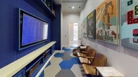 This energetic game room is thoughtfully designed to deliver an immersive entertainment experience. The most striking architectural feature is the bold, cobalt-blue built-in wall unit, which houses a large wall-mounted flat-screen television above an open shelving console complete with storage baskets and display space — ideal for gaming equipment and media accessories. The floors are a standout element, featuring a dynamic pattern of large hexagonal tiles in shades of blue, gray, and white that add visual energy and a playful, modern aesthetic throughout the space. Natural light enters through elegant glass-paned French doors, which connect the room to an adjoining area, keeping the space feeling open and bright. The neutral gray walls serve as a canvas for a curated collection of colorful, retro-style gaming and pop-culture art prints, including bold titles like "Robot Wars," "Defeat the Beast," and "Power Up," reinforcing the room's fun and spirited theme. Seating includes a row of classic wooden auditorium-style chairs and a casual leather bean bag, offering a relaxed yet character-filled atmosphere. The room flows seamlessly from the entertainment wall to the seating area, making it a well-organized and inviting space for gaming and leisure.