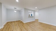 This thoughtfully renovated single-floor unit presents a clean, modern aesthetic throughout, unified by warm-toned hardwood-style flooring and soft neutral gray walls with crisp white trim. The open living and dining area is generously proportioned, with recessed lighting and a stylish multi-pendant chandelier adding visual interest overhead. A pass-through window connects this space to the kitchen, creating an open, communicative layout ideal for entertaining.

The kitchen is a standout feature, outfitted with white shaker-style cabinetry, white quartz countertops, and stainless steel appliances including a gas range with overhead microwave. A bold teal herringbone tile backsplash serves as a striking design focal point, echoed again in the bathroom, where the same tile pattern lines the tub surround from floor to ceiling. The bathroom also features large-format gray stone-look floor tile, providing a clean, spa-like finish.

The bedrooms are well-sized and bright, each with large windows framing lush green outdoor scenery. One bedroom includes a double closet with paneled doors. The living area extends toward sliding glass doors that open to an outdoor space with views of mature trees, inviting natural light deep into the interior and creating a seamless connection to the outdoors.