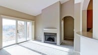 This single-floor space showcases a thoughtfully designed living area centered around a striking fireplace with dark granite surround and a clean white mantel. The vaulted ceiling above the fireplace wall creates an impressive sense of vertical space, adding to the overall openness of the room. Neutral gray-taupe walls complement the plush wall-to-wall carpeting, contributing to a cohesive and calming aesthetic throughout.

Natural light fills the living area through large sliding glass doors, which provide access to an outdoor balcony and offer views of surrounding mature trees. The doors are framed by sheer panel curtains that soften the light entering the space.

An elegant arched doorway leads from the main living area into an adjacent hallway or secondary space, adding an architectural detail that enhances the overall character of the interior. The clean lines and minimal design elements throughout give the space a move-in-ready feel, offering a versatile canvas suitable for a variety of interior styles and furnishing arrangements.