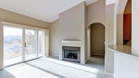 This single-floor space showcases a thoughtfully designed living area centered around a striking fireplace with dark granite surround and a clean white mantel. The vaulted ceiling above the fireplace wall creates an impressive sense of vertical space, adding to the overall openness of the room. Neutral gray-taupe walls complement the plush wall-to-wall carpeting, contributing to a cohesive and calming aesthetic throughout.

Natural light fills the living area through large sliding glass doors, which provide access to an outdoor balcony and offer views of surrounding mature trees. The doors are framed by sheer panel curtains that soften the light entering the space.

An elegant arched doorway leads from the main living area into an adjacent hallway or secondary space, adding an architectural detail that enhances the overall character of the interior. The clean lines and minimal design elements throughout give the space a move-in-ready feel, offering a versatile canvas suitable for a variety of interior styles and furnishing arrangements.