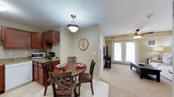 This single-floor residence offers a cohesive and inviting open-concept layout that seamlessly connects the kitchen, dining area, and living room. The kitchen is outfitted with rich, dark cherry wood cabinetry, granite countertops, and space for essential appliances, providing both functionality and visual warmth. Adjacent to the kitchen, the dining area features a classic pendant light fixture and comfortably accommodates a round dining table with upholstered seating — ideal for everyday meals or casual entertaining. The transition from tile flooring in the kitchen and dining area to plush carpeting in the living room subtly defines each zone while maintaining an open and airy feel throughout. The living room is bright and welcoming, benefiting from generous natural light that streams in through a set of elegant French doors. A ceiling fan adds comfort and practicality to the space. Neutral, warm-toned walls throughout create a versatile backdrop that complements the rich wood finishes and carpeted floors. The overall ambiance is cozy yet refined, combining traditional design elements with everyday livability in a well-connected, single-level floor plan.