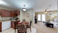 This single-floor residence offers a cohesive and inviting open-concept layout that seamlessly connects the kitchen, dining area, and living room. The kitchen is outfitted with rich, dark cherry wood cabinetry, granite countertops, and space for essential appliances, providing both functionality and visual warmth. Adjacent to the kitchen, the dining area features a classic pendant light fixture and comfortably accommodates a round dining table with upholstered seating — ideal for everyday meals or casual entertaining. The transition from tile flooring in the kitchen and dining area to plush carpeting in the living room subtly defines each zone while maintaining an open and airy feel throughout. The living room is bright and welcoming, benefiting from generous natural light that streams in through a set of elegant French doors. A ceiling fan adds comfort and practicality to the space. Neutral, warm-toned walls throughout create a versatile backdrop that complements the rich wood finishes and carpeted floors. The overall ambiance is cozy yet refined, combining traditional design elements with everyday livability in a well-connected, single-level floor plan.