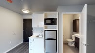 This single-floor studio unit offers a thoughtfully organized layout that maximizes every square foot. The kitchenette is outfitted with white cabinetry, a clean white countertop with an undermount sink, a compact refrigerator, a microwave, and open shelving — providing all the essentials for day-to-day living in a streamlined footprint. The cabinetry features sleek, modern hardware, and the white surfaces contrast cleanly against the dark wood-style flooring that runs throughout the main area. A dark-finished entry door adds a bold design accent at the unit entrance. The bathroom, visible through an open doorway, is finished in neutral tones with a toilet and appears to include a grab bar, suggesting accessibility-conscious design. Flush-mounted ceiling lighting keeps the space bright and well-lit. The overall color palette — soft grays, whites, and dark wood accents — creates a clean, contemporary aesthetic. This unit is well-suited for those seeking a functional, low-maintenance living space with modern conveniences efficiently integrated into a compact design.